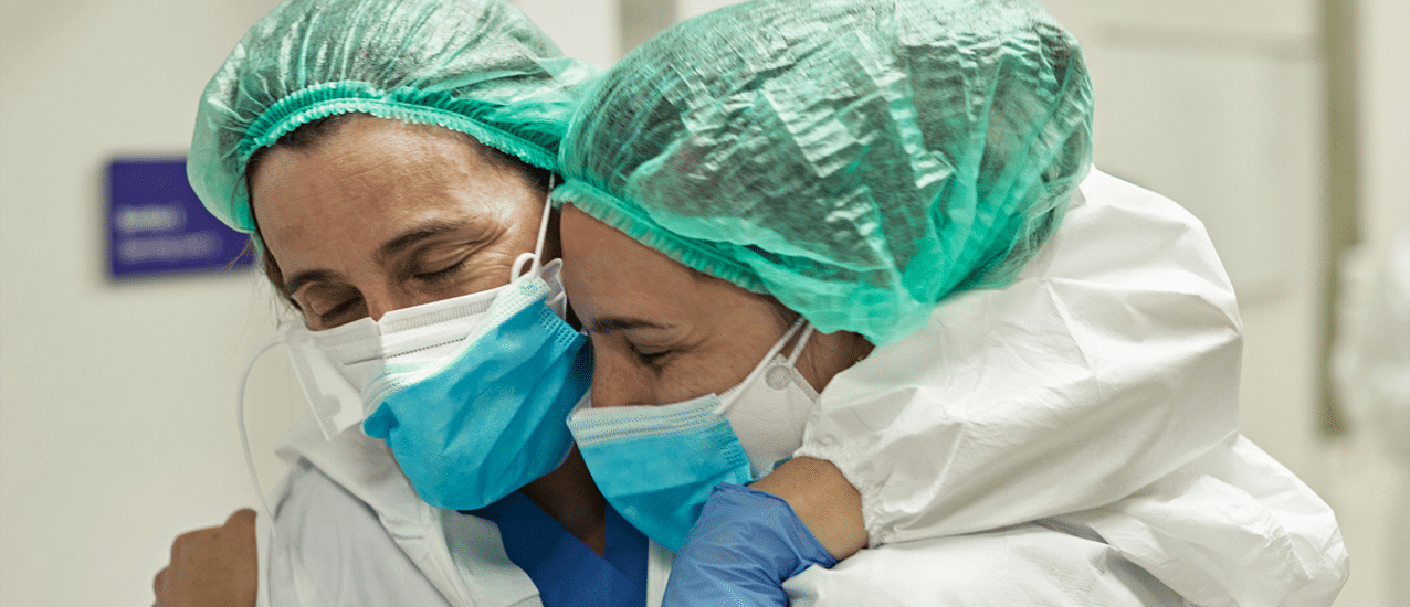 Two healthcare workers wearing masks, gloves and, and other protective gear embrace in a hug.
