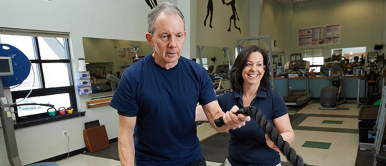 A smiling female physical therapist helps a mature male patient perform a rope exercise in a gym.