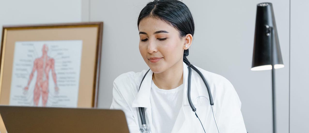 A young female physician seated at an office desk working on a laptop computer.