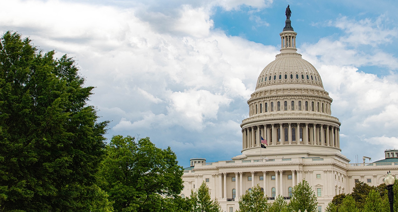 A view of the United States Capitol building surrounded by trees and a cloudy blue sky.