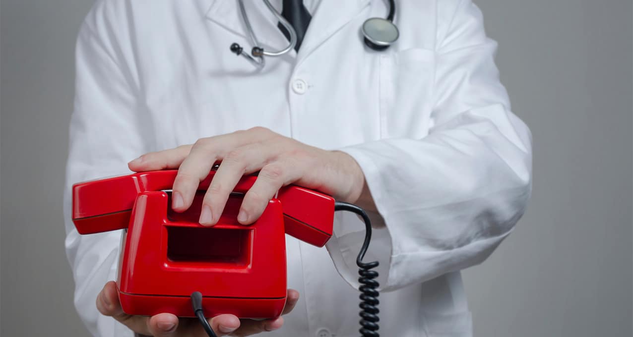Close-up of a doctor holding a red landline telephone with one hand resting on the handset.